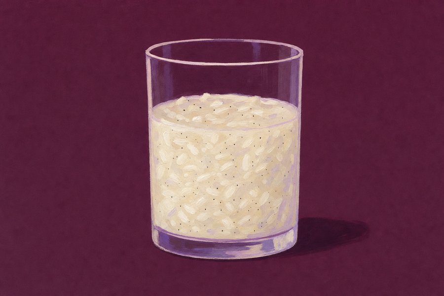A plain straight-sided glass two-thirds filled with loose creamy rice pudding, individual swollen grains visible and flecked with dark vanilla seeds.